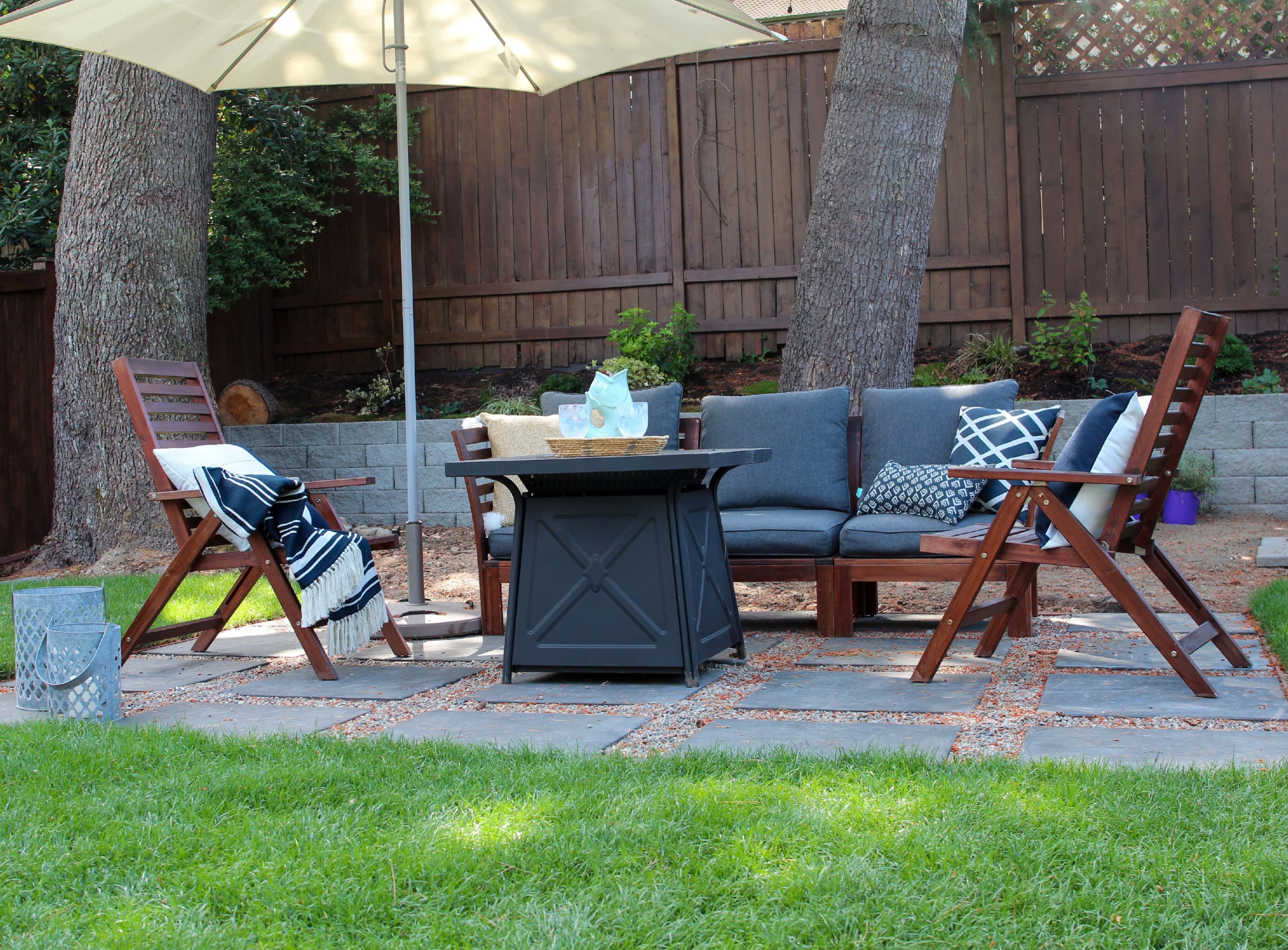 Patio of dark gray pavers and gravel in between. Brown wood patio furniture with gray cushions and navy blue accent pillows. Black metal fire pit table and umbrella.